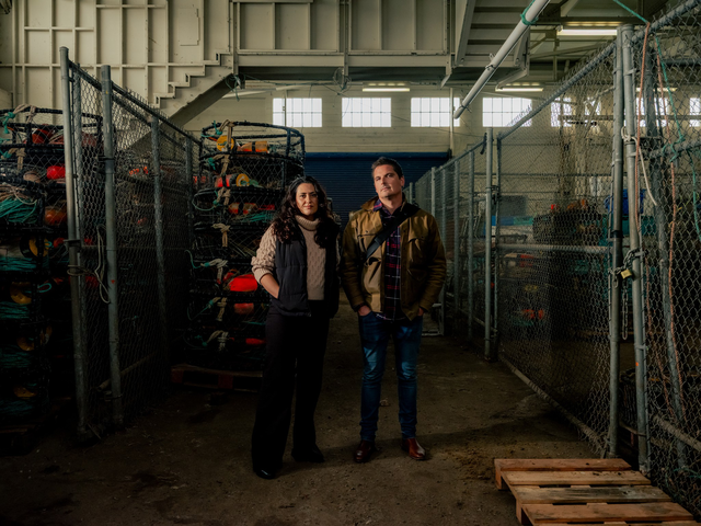 A man and woman stand side by side in a dimly lit storage area with metal fencing and stacked equipment cases around them.